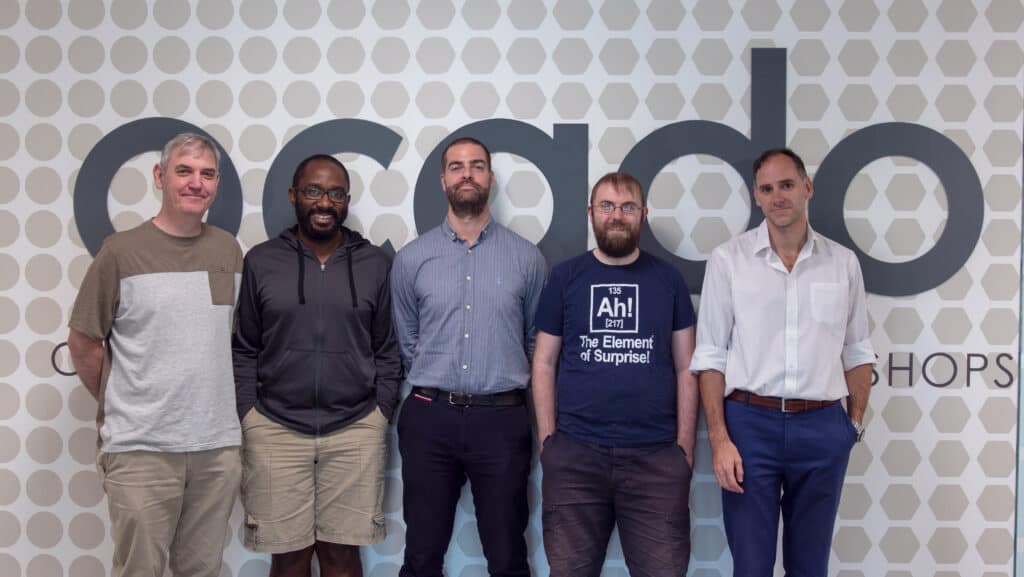 Five gentlemen taking picture in front of ocado wallpaper