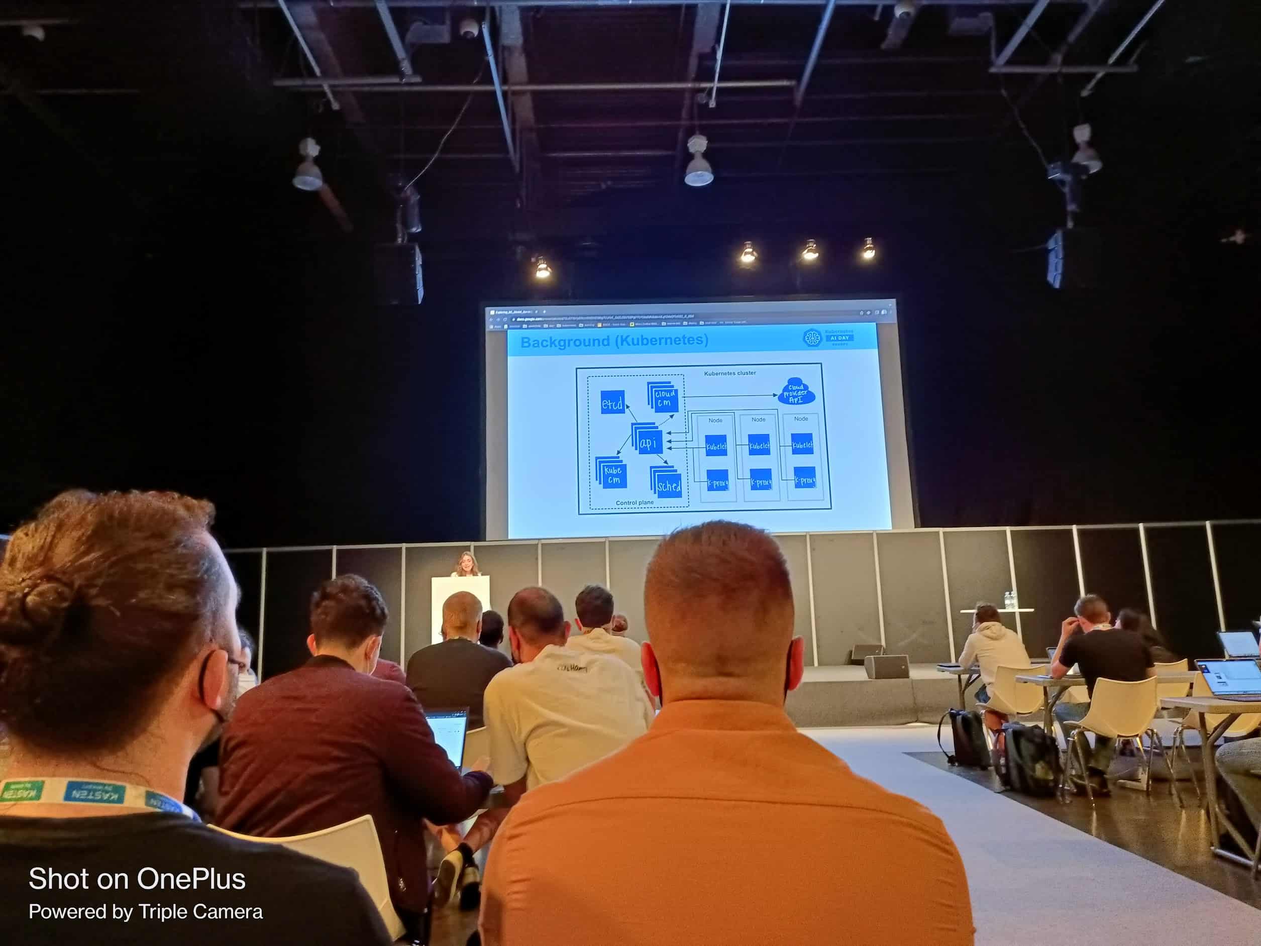 A lady presenting Background (Kubernetes) in front off audience with laptop in the hall