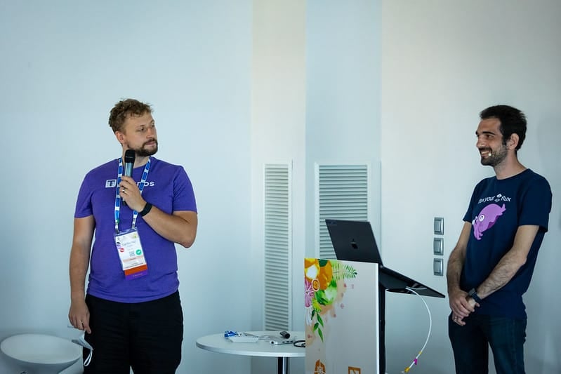 Stefan Prodan smiles to audience accompanied by a gentleman holding a microphone in meeting hall