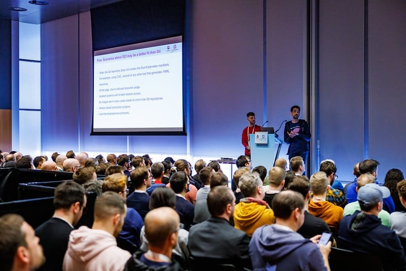 Two gentlemen giving presentation on the stage in front of participants in the hall regarding "Flux Scenarios where CCI may be better fit than Git"