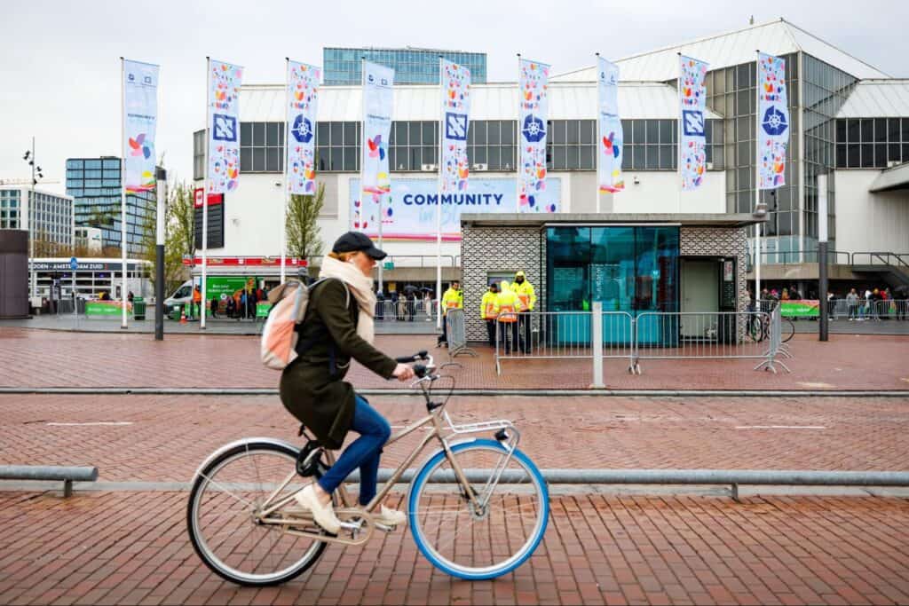 A lady cycling in front of KubeCon hall with hat, bag and headset