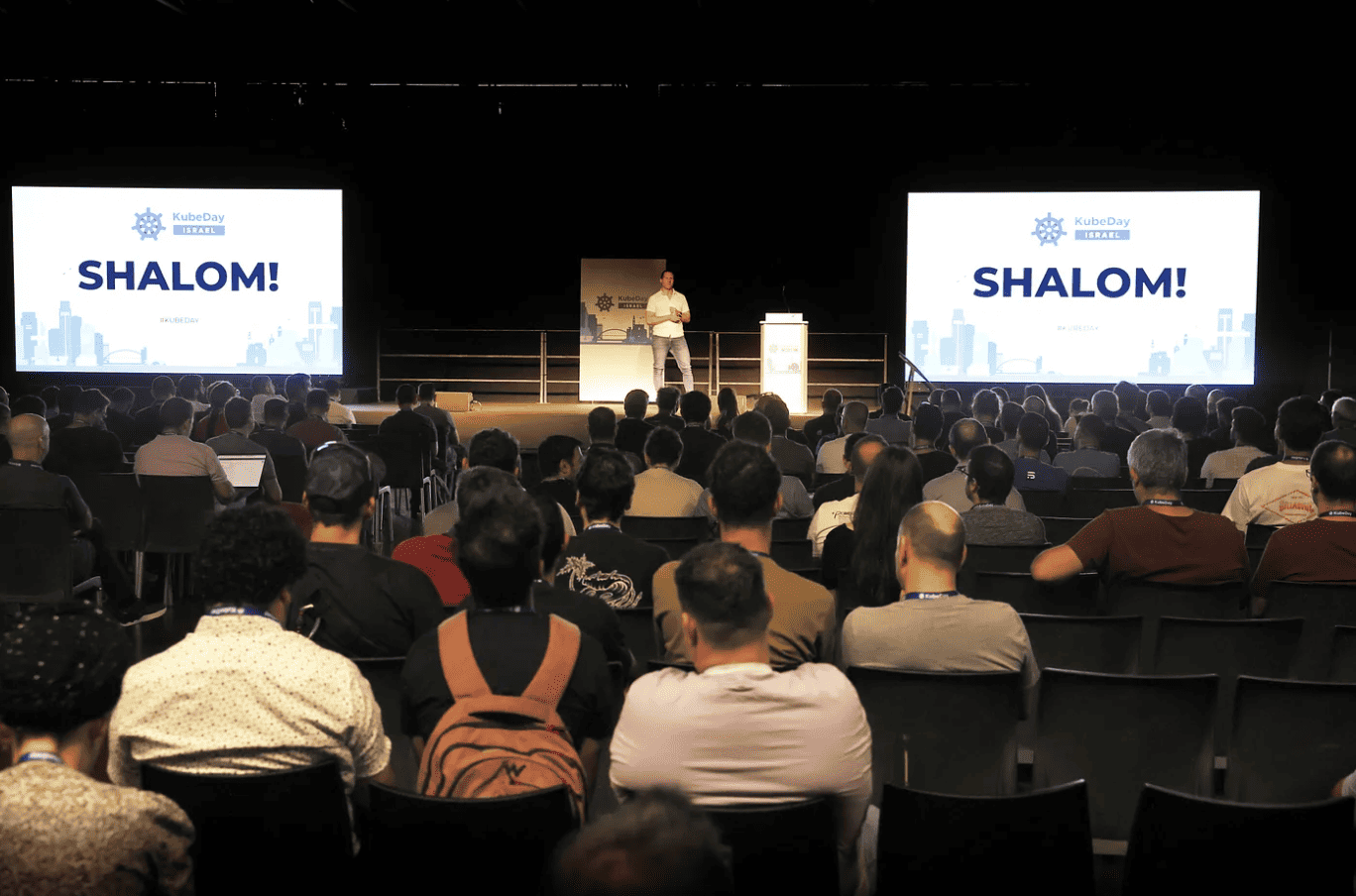 A gentleman talking on the stage on KubeDay Israel with 2 projectors saying "Shalom"