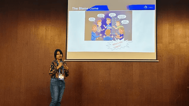 A photo image of 'The Blame Game' presentation slide on a projector screen, against a bronze wall backdrop, with the presenter in the foreground at KCD Sri Lanka 2025.