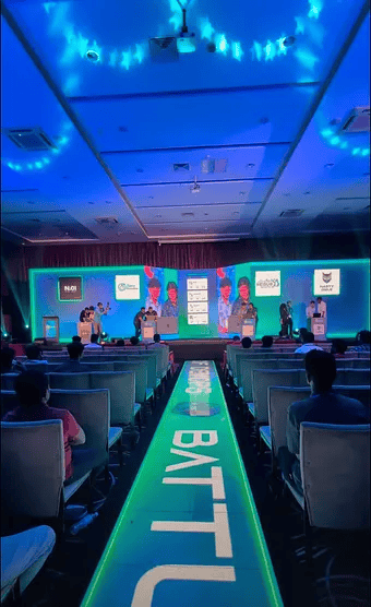 A photo image of 'Battle of DevOps' at KCD Sri Lanka 2025. The image shows the auditorium room and stage area lit up with green and blue lighting, and a green neon aisle and seating area. 