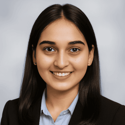 A profile portrait of Nupur wearing a black jacket and blue buttoned shirt, against a grey backdrop. Nupur is smiling.  