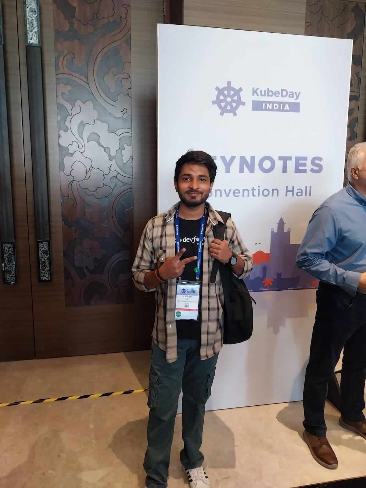 Mentee Lavish Pal, wearing a brown check shirt and black t-shirt, with a blue lanyard around his neck, standing in front of a KubeDay India advertisement, smiling into the camera. 