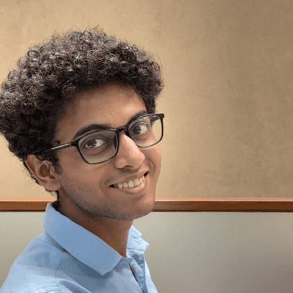 A side profile photo of Rishab, smiling and wearing glasses and a blue buttoned shirt. He is against a beige and wood panelled backdrop. 