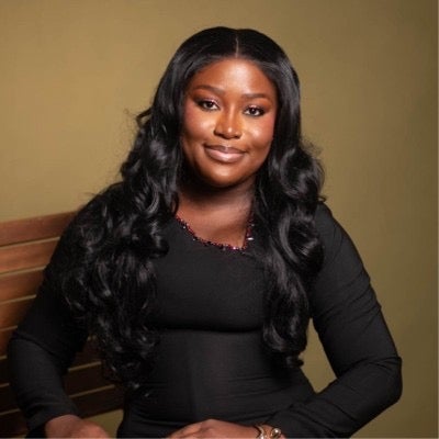 A photo of Peace Sandy, sitting in a chair with her arms crossed. wearing a black dress. Peace is smiling. She is against a gold backdrop. 