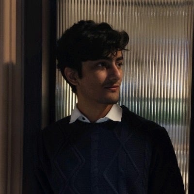 A side profile image of Shivang wearing a black jumper and white shirt, against a glass backdrop. 