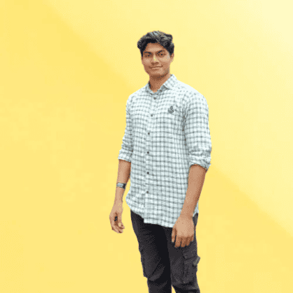 A full standing shot of Saurav, wearing a white and green check shirt and grey jeans, standing against a yellow backdrop. 
