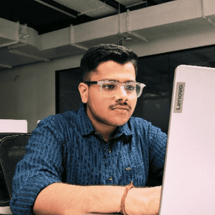 An image of Yash, wearing glasses and a blue stripe shirt, sitting at a desk and working at a Lenovo desktop computer. 