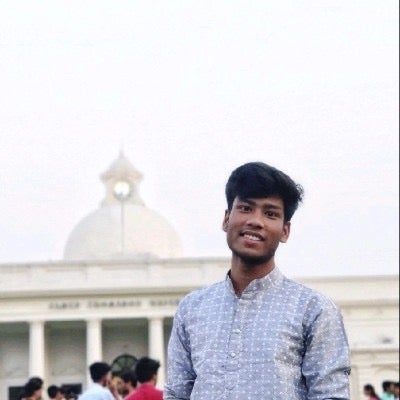 Mentee Adesh Pal standing outside a white building smiling, wearing a light blue and white collarless shirt, with a crowd of people behind him. 