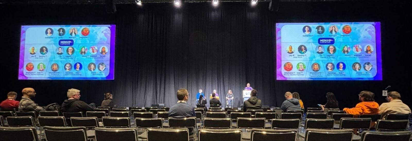 A photo of the Merge Forward panel discussion - with 3 panelists seated and one moderator standing on stage, between two screens. The photo also features the audience. 