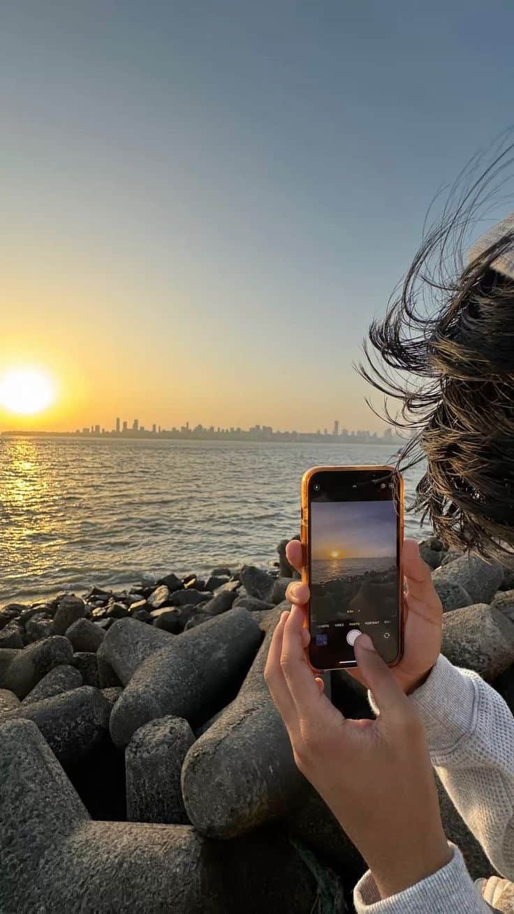 A photo of one of the authors using their iPhone to take a photo of the sunset over the sea and city skyline. There are coastal rocks in the forefront of the image. 